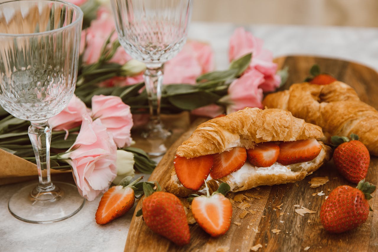 Home Close-up of delicious croissants with strawberries and cream, accompanied by pink flowers and wine glasses.
